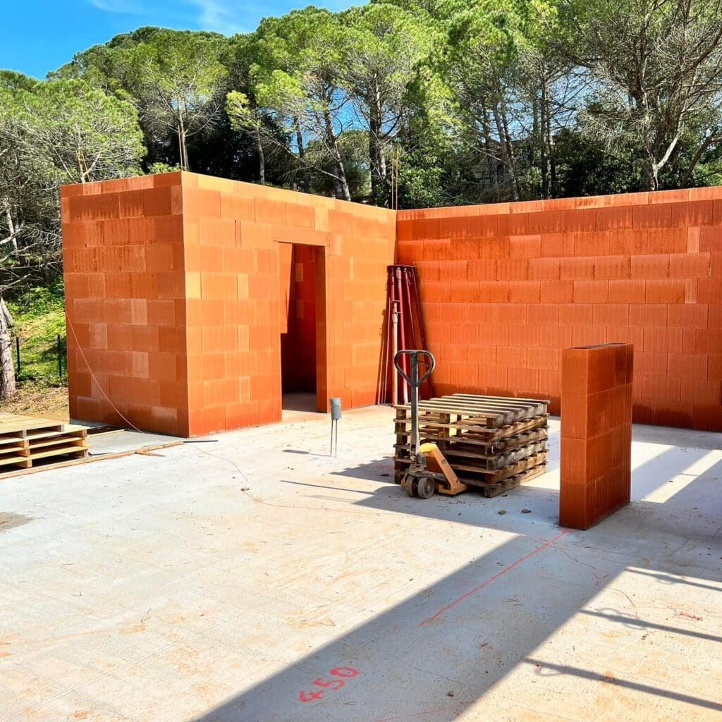 Murs en briques terre cuite : avancement de la construction Élévation des murs en briques terre cuite sur une dalle de chantier ensoleillée. Transpalette, palettes en bois et marquage au sol 450.