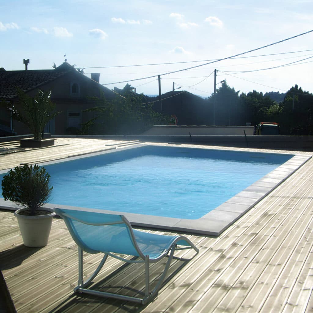 Piscine extérieure avec terrasse en bois et transat bleu. Vue sur une piscine extérieure entourée d'une terrasse en bois et d'un transat bleu. Fond de ciel bleu, maisons et arbres sombres.