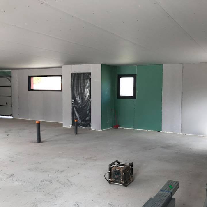 Chantier isolation : Pose de placo hydrofuge et cloisons. Intérieur de chantier : pose de cloisons en placo hydrofuge vert et gris. Porte bâchée, deux fenêtres noires, sol en béton et tuyaux d'attente.